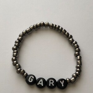 Pu&ograve; includere: Un braccialetto di perline argentate con perline a forma di lettera nere che compongono il nome "GARY".