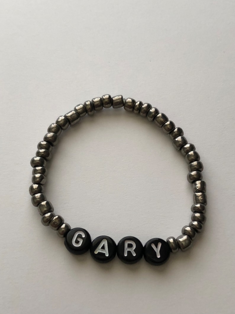 Pu&ograve; includere: Un braccialetto di perline argentate con perline a forma di lettera nere che compongono il nome "GARY".