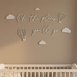 Op de afbeelding: Een kinderkamerscène met een witte wieg en beddengoed, een dressoir en wanddecoraties. De wandkunst bevat de zin "Oh the places you'll go" met luchtballonnen en wolken. Rechts is een raam met witte gordijnen.
