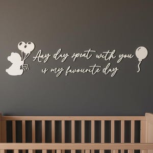May include: White wooden wall art with the phrase "Any day spent with you is my favourite day." The art features a bear holding heart-shaped balloons and a single balloon. A light-coloured wooden cot is in the foreground.
