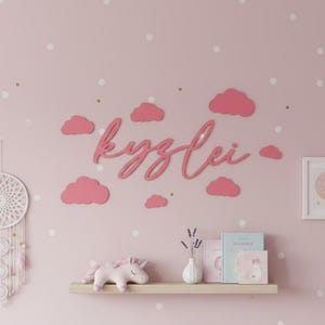 May include: A pink wall with white polka dots features a dreamcatcher, a cursive name, and pink cloud cutouts. A shelf holds a unicorn, books, and a vase. A framed picture of a crescent moon and stars is also present.