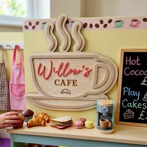 May include: Wooden play cafe sign with the text "Willow's Cafe" and a coffee cup design. A small chalkboard lists "Hot Cocoa" and "Play Cakes." Toy food items include a muffin, croissant, sandwich, and macarons.