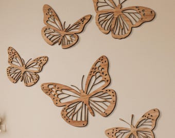 May include: Five wooden butterfly wall decorations in various sizes are arranged on a beige wall. The butterflies are cut from light brown wood, with intricate wing patterns. The butterflies are a decorative accent.