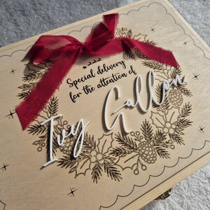 May include: A rectangular wooden box featuring a laser-engraved wreath design with pine needles and pinecones. A dark red fabric ribbon is tied at the top. The text reads: Special delivery for the attention of Ivy Gallon in white script.