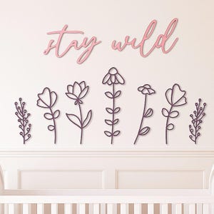 May include: A wall decoration with the words "stay wild" in pink script above a row of dark purple floral cutouts. The flowers are in various shapes and styles, creating a whimsical and natural aesthetic.