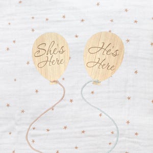 May include: Two wooden balloon-shaped decorations with the phrases "She's Here" and "He's Here" engraved on them. The balloons are attached to strings, one pink and one blue, against a white background with star patterns.