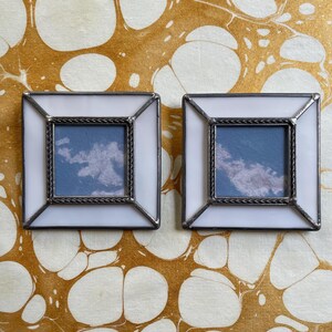 May include: Two square, white-framed miniature artworks. Each features a blue sky with clouds, framed by a silver-colored rope-like border. The frames are set against a marbled background with white and gold tones.