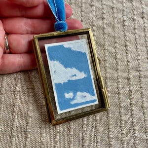 May include: A small, rectangular brass frame with a miniature painting of a blue sky with white clouds. A blue velvet ribbon is attached to the top. The frame is held against a neutral-toned fabric background.