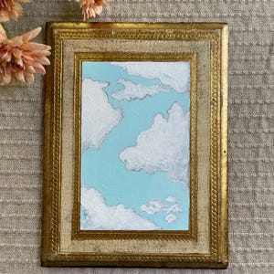 May include: A framed painting of a blue sky with fluffy white clouds. The painting is in a gold-colored ornate frame. The artwork evokes a sense of calm and serenity.