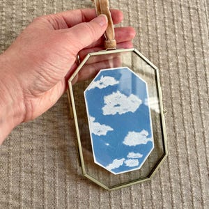 May include: A decorative glass frame with a gold-colored metal border. Inside is a blue and white cloud design. A tan velvet ribbon is attached for hanging. The frame is held by a hand.