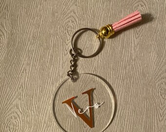 Op de afbeelding: Een transparante acryl sleutelhanger met een gouden metalen ring en een roze kwast. De sleutelhanger heeft een gouden letter "V" met de naam "Vera" in wit schrift.