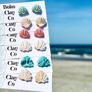 May include: Six pairs of seashell-shaped stud earrings displayed on white cards labeled Boho Clay Co. The earrings feature various colors and textures, including solid mint, beige, coral, speckled white, marbled teal, and marbled tan clay finishes.