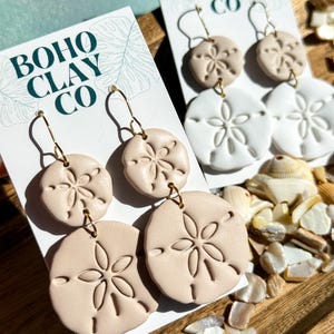 May include: Sand dollar-shaped earrings in beige and white, displayed on cards with the text "BOHO CLAY CO." The earrings have gold-colored hooks and are set against a wooden tray with seashells.
