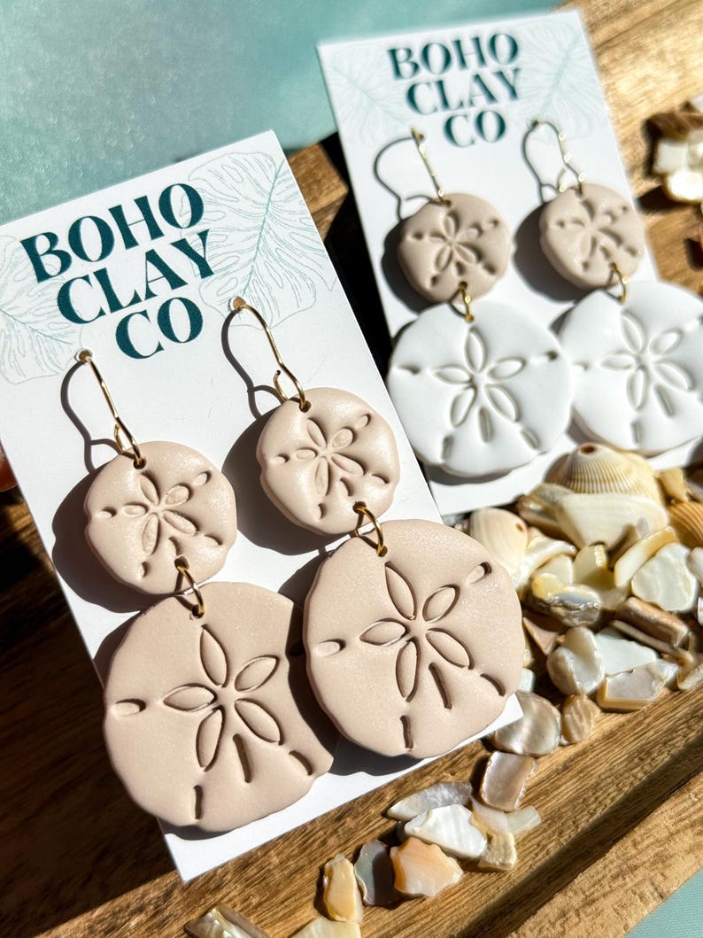 May include: Sand dollar-shaped earrings in beige and white, displayed on cards with the text "BOHO CLAY CO." The earrings have gold-colored hooks and are set against a wooden tray with seashells.