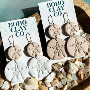 May include: Sand dollar-shaped earrings in white and beige, displayed on cards that read "BOHO CLAY CO." The earrings are on gold-colored hooks. The background includes seashells and decorative stones, creating a beachy aesthetic.