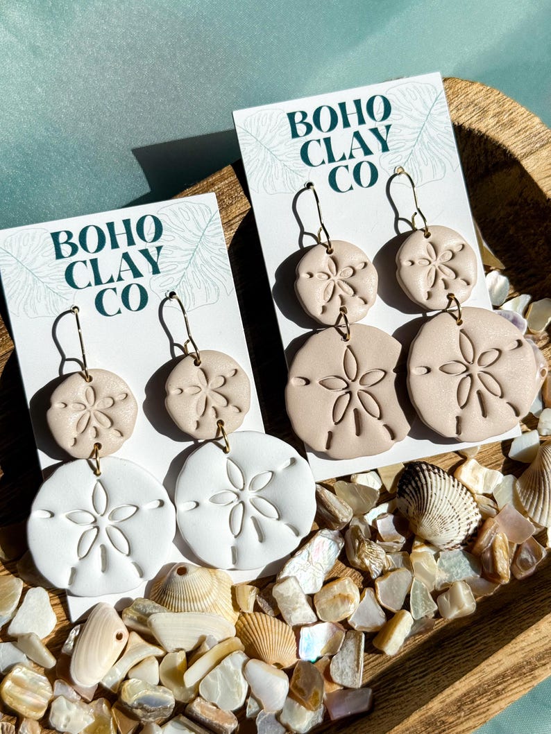 May include: Sand dollar-shaped earrings in white and beige, displayed on cards that read "BOHO CLAY CO." The earrings are on gold-colored hooks. The background includes seashells and decorative stones, creating a beachy aesthetic.