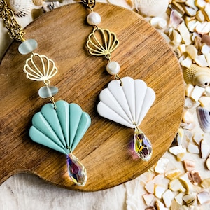 May include: Two necklaces featuring gold-tone chains, white pearl beads, and gold-tone shell charms. One necklace has a light green shell pendant and frosted glass beads, while the other has a white shell pendant and a faceted iridescent crystal drop.