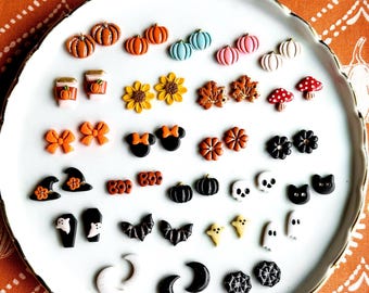 May include: A collection of small stud earrings arranged on a white plate, featuring various shapes including pumpkins, sunflowers, leaves, mushrooms, bows, mouse ears, witch hats, skulls, cats, ghosts, bats, moons, and spider webs in diverse colors.