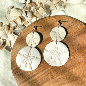 May include: Cream-colored, double-drop earrings featuring a sand dollar design. The earrings have a gold-colored post and connecting hardware. The sand dollar shapes are detailed with etched patterns. The earrings are displayed on a wooden surface.