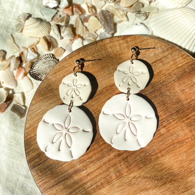 May include: Cream-colored, double-drop earrings featuring a sand dollar design. The earrings have a gold-colored post and connecting hardware. The sand dollar shapes are detailed with etched patterns. The earrings are displayed on a wooden surface.
