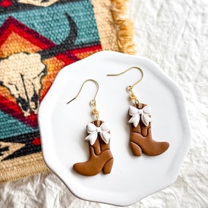 May include: A pair of brown cowboy boot earrings with white bows. The earrings are on a white plate with a gold background.