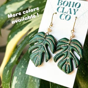 May include: A pair of dark green monstera leaf earrings with gold glitter accents in the leaf veins. The earrings feature small white beads and gold-toned metal hooks. They are attached to a white display card labeled BOHO CLAY CO.