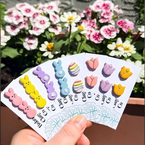 May include: A hand holds a fan of colorful clay Easter-themed decorations. The decorations include pink, yellow, and purple bunnies, striped eggs, and tulips in pink, purple, and yellow. The text "Boho Clay Co" is visible.
