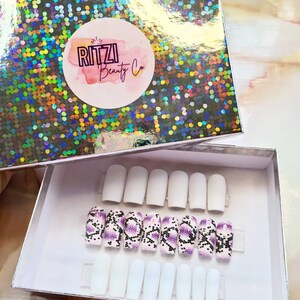 May include: A white box with a purple and black snake skin design on the nails inside. The box has the logo "RITZI Beauty Co." on the lid.