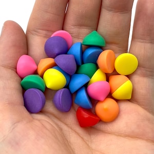 May include: A handful of colorful, small, geometric-shaped objects in various colors including pink, purple, green, yellow, orange, and blue. The objects are held in an open hand against a white background.