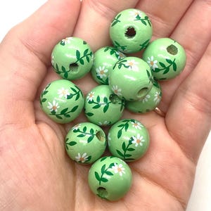 May include: A collection of green wooden beads with white flowers and green leaves painted on them. The beads have a hole in the center for stringing.