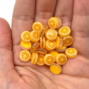May include: A handful of miniature orange slices, featuring a bright orange interior with white segments and a yellow rind. The small, round pieces are translucent and appear to be made of resin or a similar material. The overall theme is food-related.