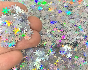 May include: A pile of iridescent silver snowflake glitter. The glitter is holographic and reflects rainbow colors.