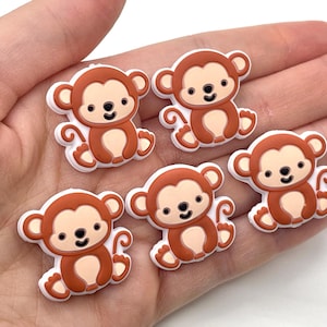 May include: Five brown and white monkey-shaped charms. Each charm features a smiling monkey with a light brown body, a darker brown head, and white accents. The charms are designed with a cartoonish style.