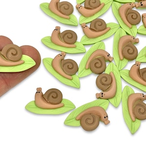 May include: A collection of brown snail charms on green leaves. The snails have a brown shell with a spiral design and a brown body with a small face. The leaves are a light green color and have a smooth surface.