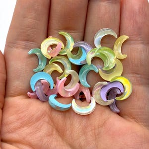 May include: A collection of colorful, crescent-shaped beads in shades of yellow, pink, blue, and purple. The beads are translucent and appear to be made of plastic or resin. They are held in a hand.