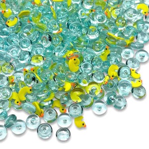 May include: A collection of clear, round glass beads with a light blue tint, interspersed with yellow duck-shaped charms. The beads vary in size and are scattered across a white surface, creating a playful and whimsical aesthetic.