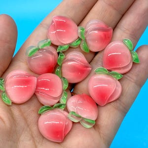 May include: A collection of small, smooth, peach-shaped cabochons featuring a gradient of pink and white colors with translucent green leaves attached to the top of each piece. These decorative items have a glossy finish and rounded shape.