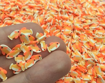 May include: A handful of small, orange, white, and yellow fish-shaped polymer clay pieces. The fish have black eyes and are scattered across a surface, creating a vibrant, colorful display. These decorative pieces are ideal for crafting and embellishments.