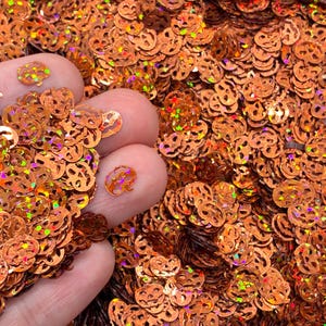 May include: A collection of small, orange, pumpkin-shaped sequins with a holographic finish. Each individual piece features a cut-out jack-o'-lantern face design. The sequins are metallic and reflective, creating a shimmering effect across the pile.