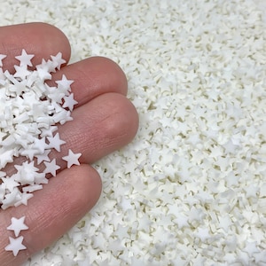 May include: A close-up of a hand holding a pile of white star-shaped sprinkles. The background is filled with more white star sprinkles.