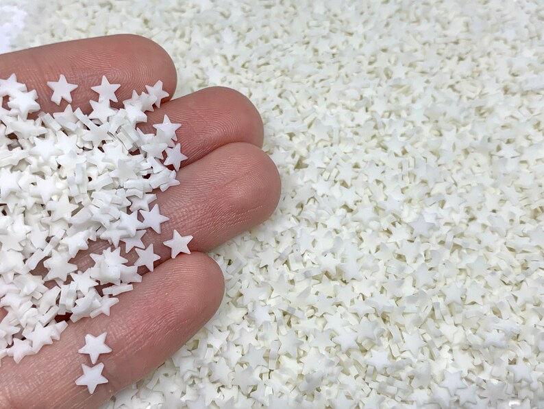 May include: A close-up of a hand holding a pile of white star-shaped sprinkles. The background is filled with more white star sprinkles.