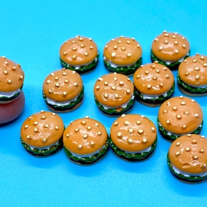 May include: Miniature burger charms with brown buns and white and green fillings are arranged on a bright blue surface. The tiny burgers have small, gold-colored dots on the top bun. One burger is held by a finger.