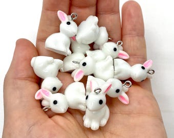 May include: A handful of small, white bunny charms with pink inner ears and black eyes. Each charm has a silver-colored loop at the top for attaching to jewelry or other crafts. The bunnies are in various poses, some sitting and some standing.