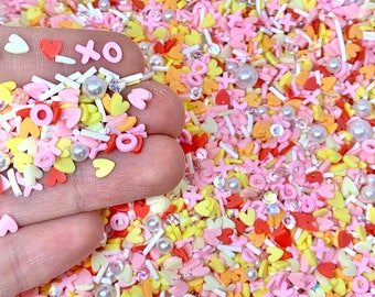 May include: A mix of colorful polymer clay sprinkles in various shapes, including hearts, flowers, and the letters "X" and "O". The sprinkles are in shades of pink, yellow, orange, and red, with white accents and pearl beads. Ideal for crafting and decorating.