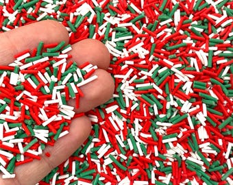 May include: Red, white, and green sprinkles in the shape of long, thin cylinders. The sprinkles are scattered on a surface and a hand is holding some of the sprinkles.