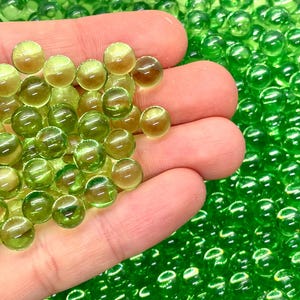 May include: A handful of translucent, round, green beads with a slight yellow hue. The beads are scattered across a surface, with some held in a hand. The beads are a decorative element, possibly for crafts or home decor.