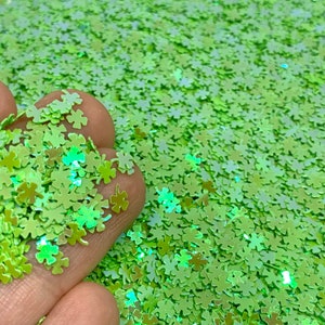 May include: Green glitter in the shape of four-leaf clovers. The glitter is iridescent and has a light green color.