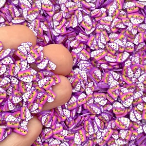 May include: A close-up of a pile of purple and pink cake slice shaped sprinkles with white frosting and a smiley face.