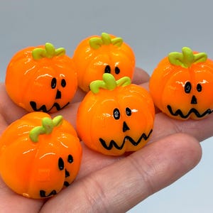 May include: Five small, bright orange pumpkin-shaped charms with glossy finishes. Each pumpkin features a light green stem at the top and a hand-painted black face with two oval eyes, a triangular nose, and a jagged, stitched-style mouth design.