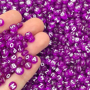 May include: A close-up of a hand holding a pile of purple alphabet beads. The beads are made of plastic and have white letters on them.